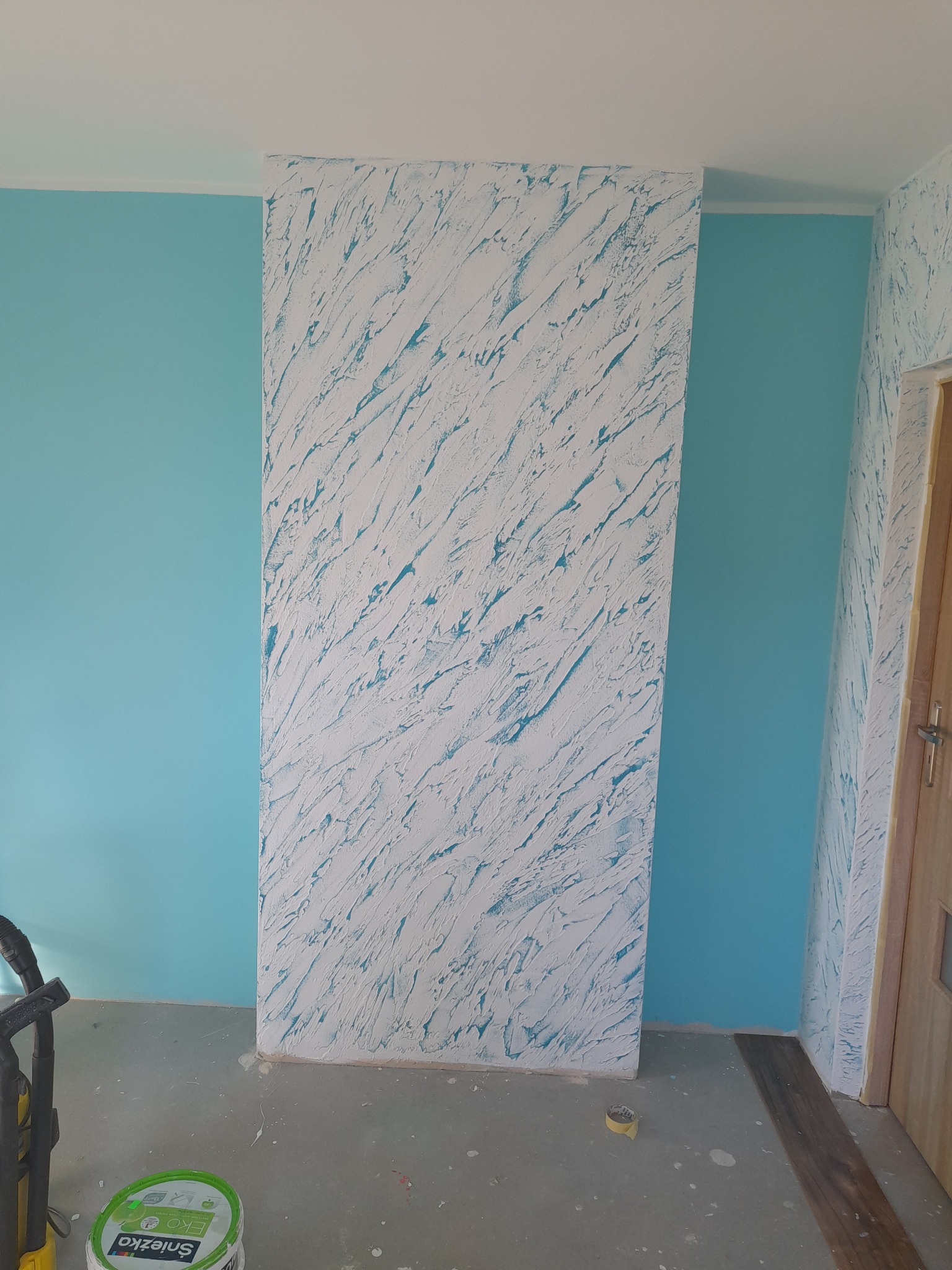 Ściana z dekoracyjną, białą strukturą i turkusowym podmalowaniem, sąsiadująca z gładką, turkusową ścianą. Widoczny fragment podłogi i drzwi.