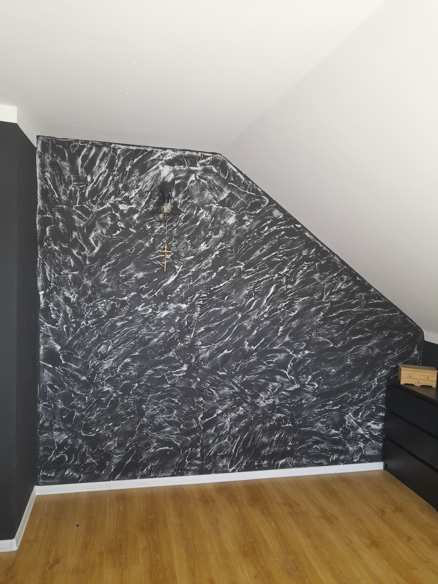 Ściana w pokoju na poddaszu, wykończona dekoracyjną, czarno-białą strukturą, z widoczną lampą wiszącą. Podłoga z paneli, biała listwa przypodłogowa.