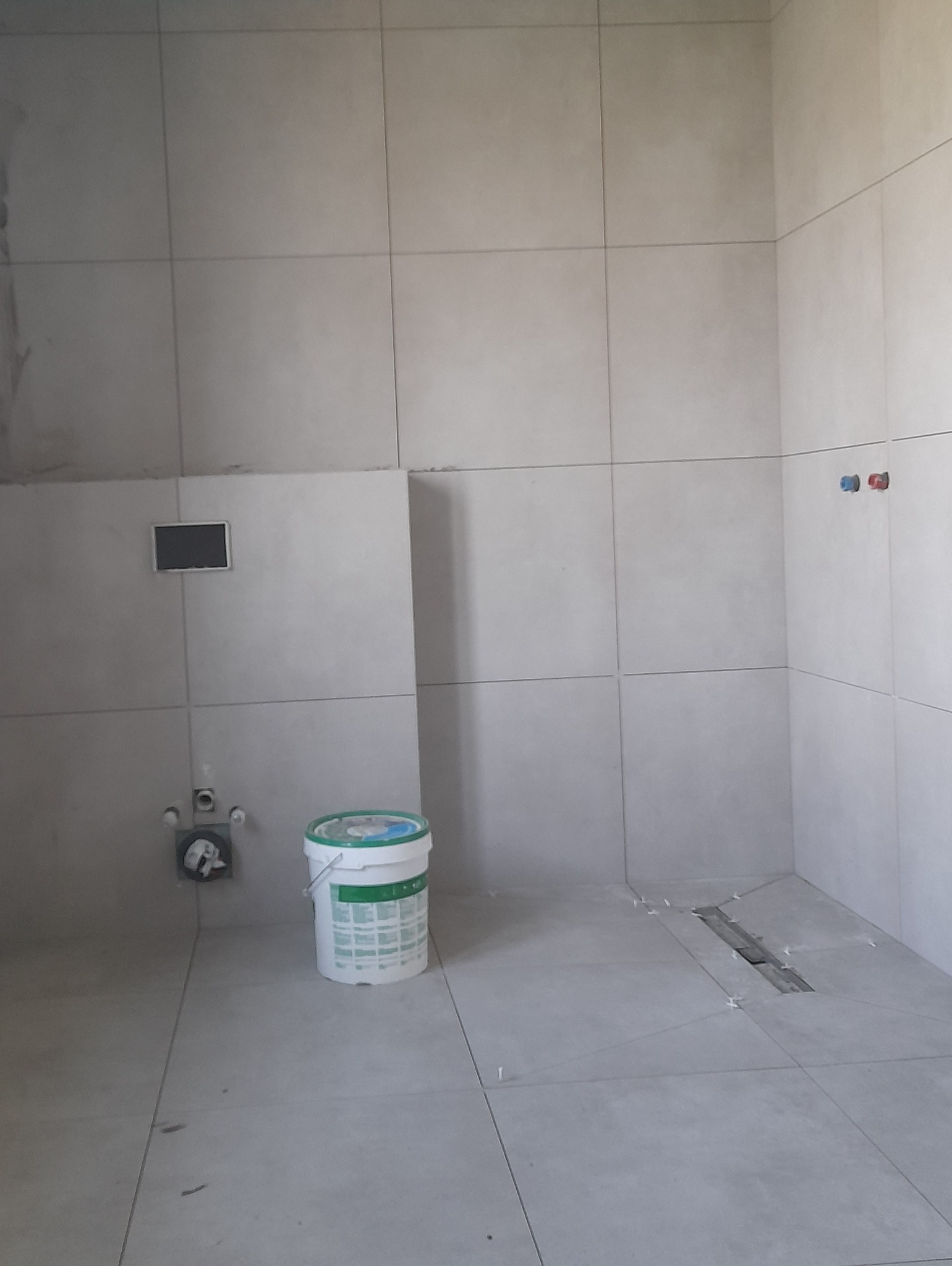 Szara łazienka w trakcie remontu: ułożone płytki, widoczny odpływ liniowy, wiadro farby. Minimalistyczny design, duże formaty płytek, etap prac wykończeniowych.