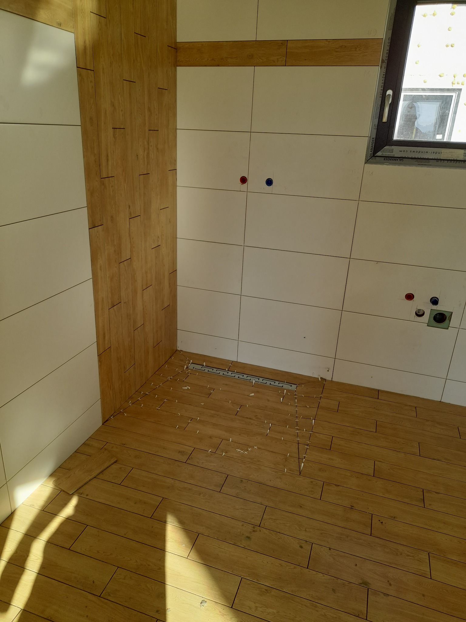 Układanie płytek imitujących drewno w łazience z odpływem liniowym. Częściowo wykończone ściany w jasnych płytkach. Widoczne krzyżyki dystansowe.