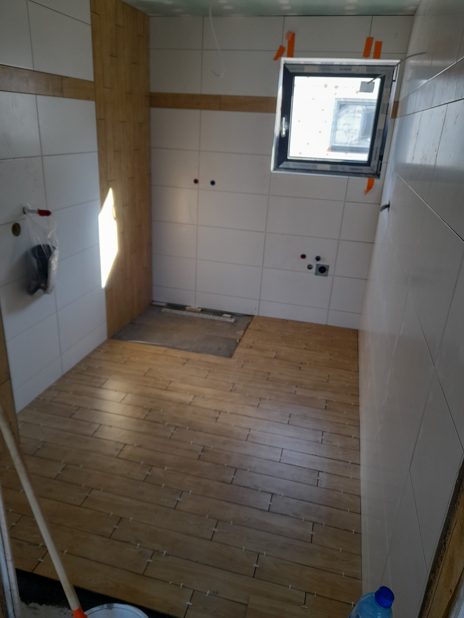 Wnętrze łazienki w trakcie wykończenia: ułożona podłoga z płytek imitujących drewno, białe płytki na ścianach, okno z pomarańczowymi taśmami. Widoczne narzędzia pracy.