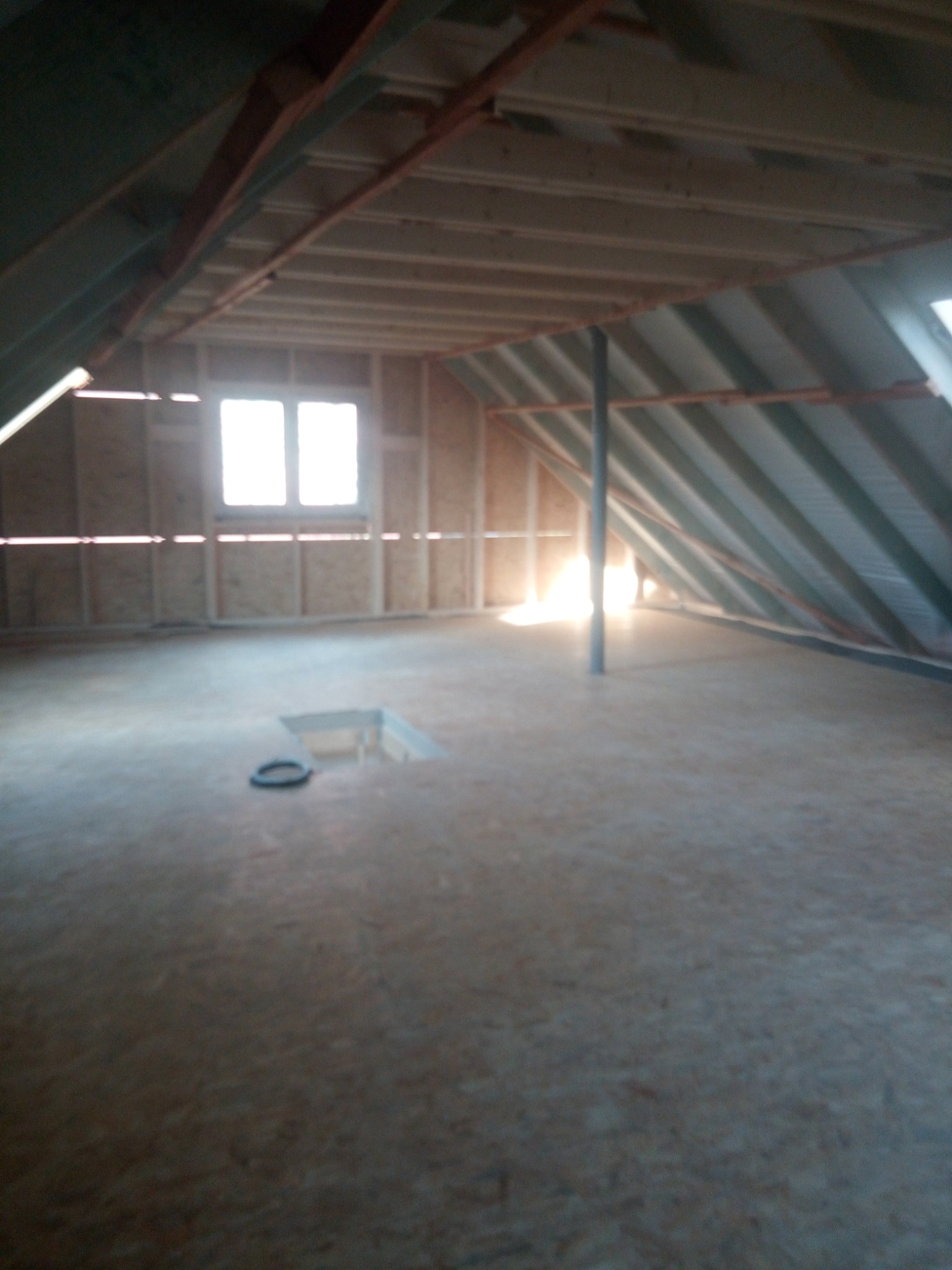 Poddasze w trakcie adaptacji w Łącku. Widoczne drewniane elementy konstrukcyjne, okno i otwór w podłodze. Etap prac wykończeniowych.