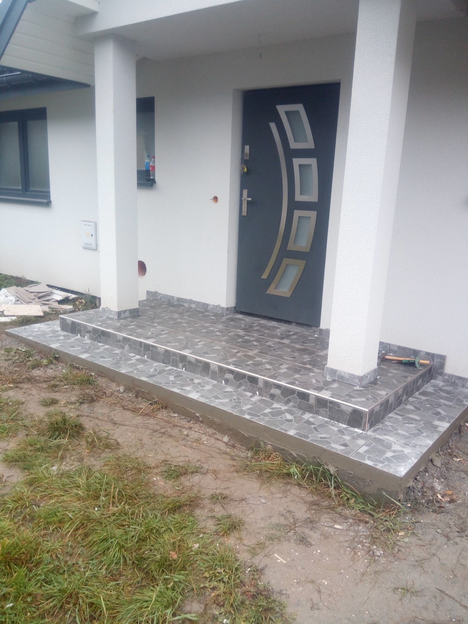 Wykończone płytkami schody wejściowe do domu z szarą, nowoczesną stolarką drzwiową. Widoczne narzędzia glazurnicze i fragmenty płytek obok schodów.