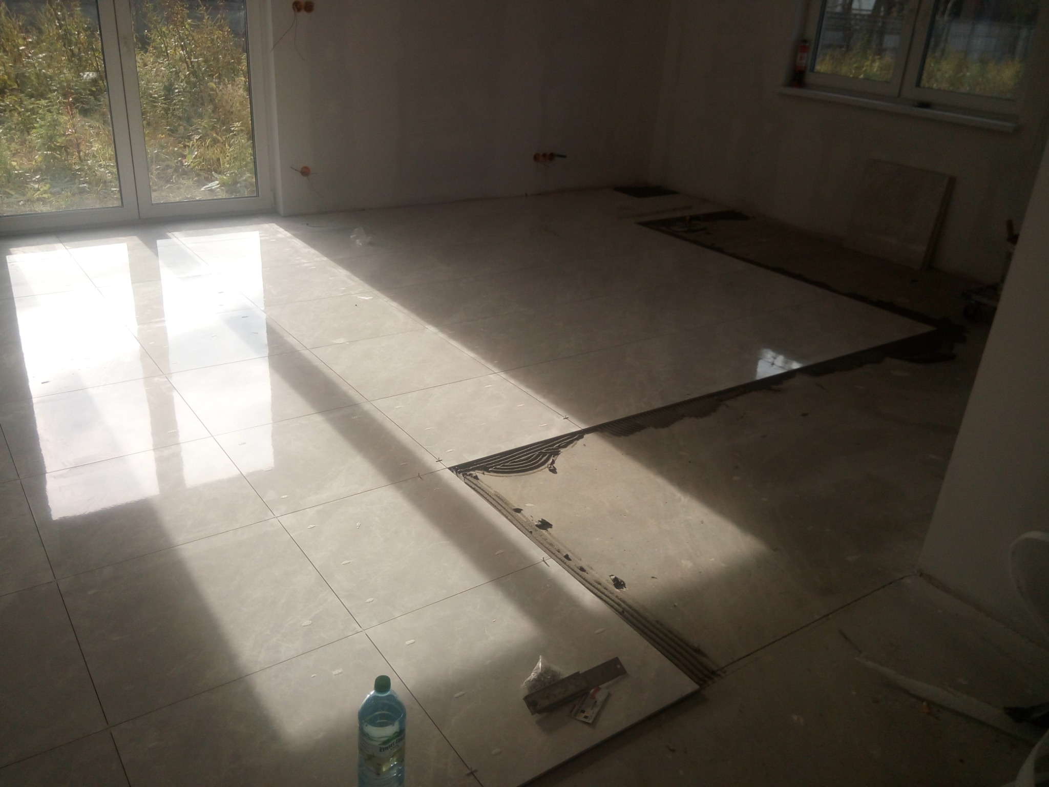 Układanie jasnych, błyszczących płytek podłogowych w nowym pomieszczeniu. Częściowo zakończone, widoczne narzędzia glazurnicze i butelka wody. Duże okno w tle.