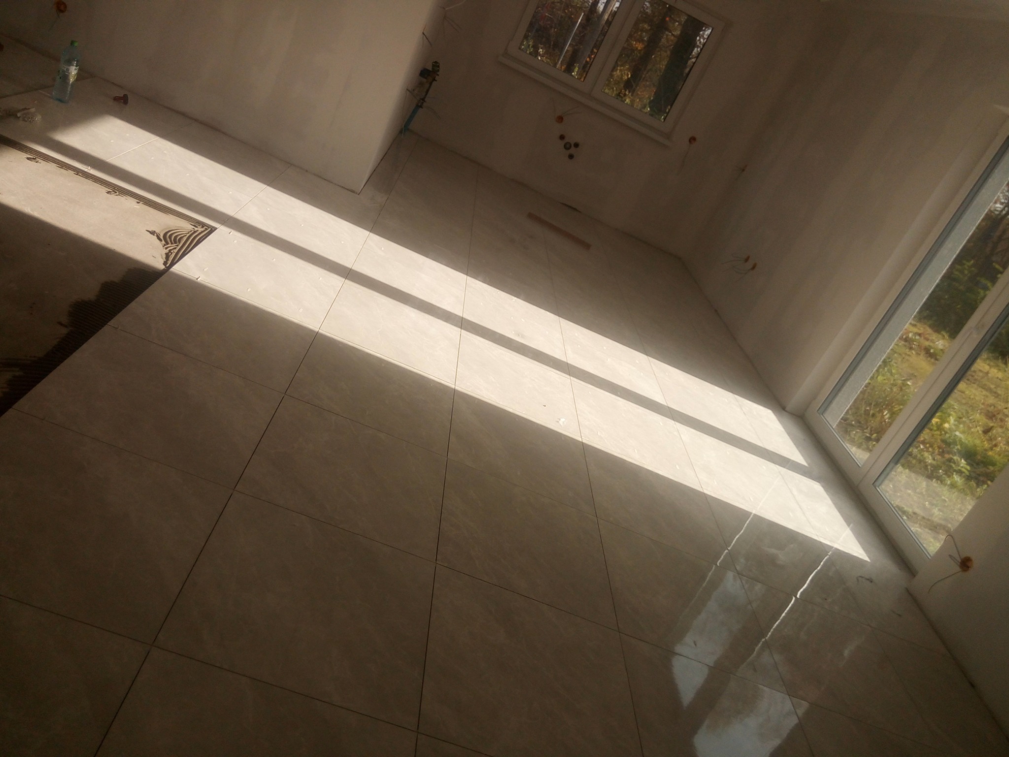 Jasne, połyskujące płytki podłogowe w nowo wykończonym wnętrzu z oknami, wpadające promienie słoneczne. Częściowo ułożona podłoga, widoczne przygotowanie podłoża.