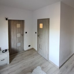 Jacek Klimek - Wnętrze po remoncie: dwie drewniane drzwi z przeszkleniami, jasne ściany i podłoga z paneli. Widoczny fragment szafki nocnej i czujnik na ścianie. Minimalistyczny design.