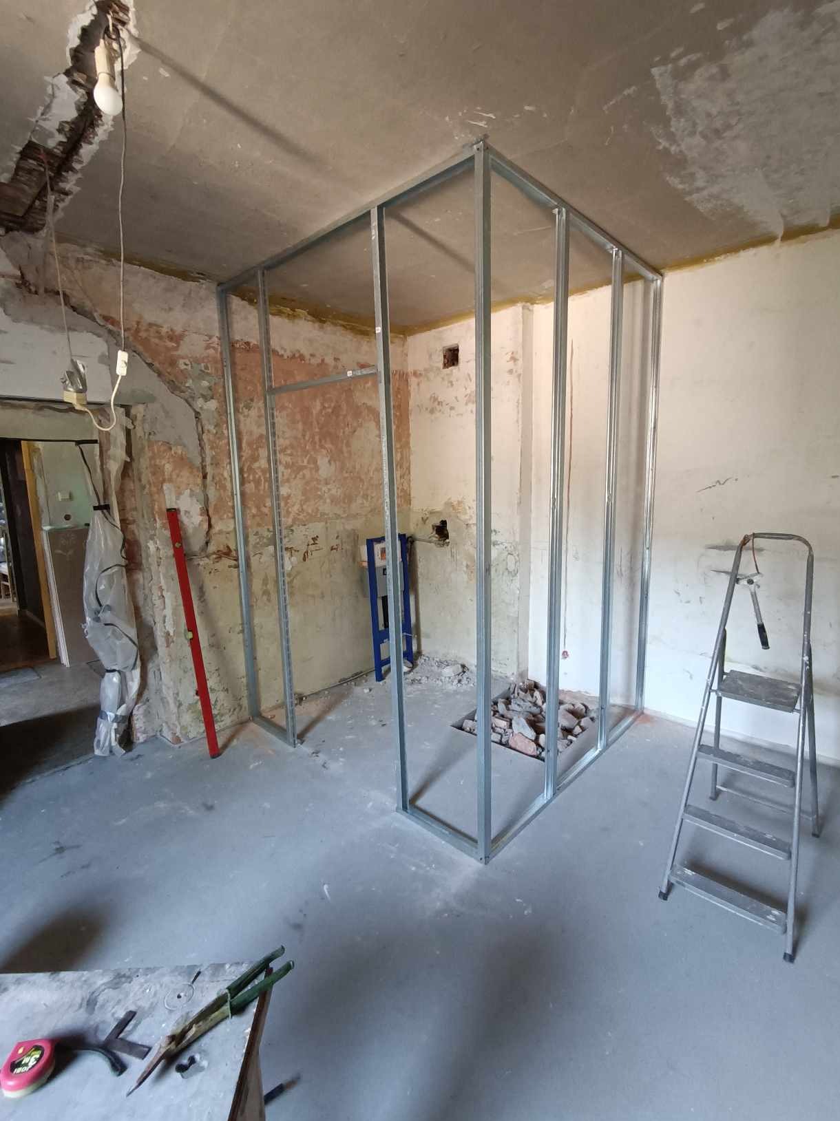 Wnętrze w trakcie remontu: metalowa konstrukcja ścianki działowej na tle surowych ścian i podłogi. Widoczne narzędzia i drabina.