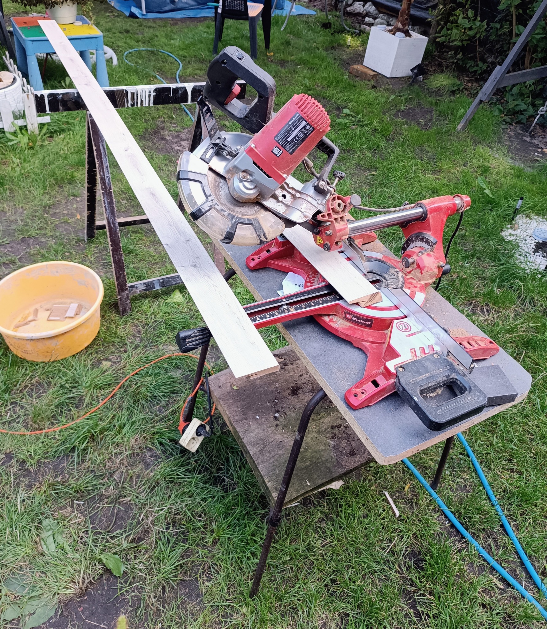Czerwona piła ukośnica Einhell tnie deskę na trawie. Przygotowanie do montażu podłogi. Deska z jasnego drewna leży na stojaku. Widoczne narzędzia i akcesoria.