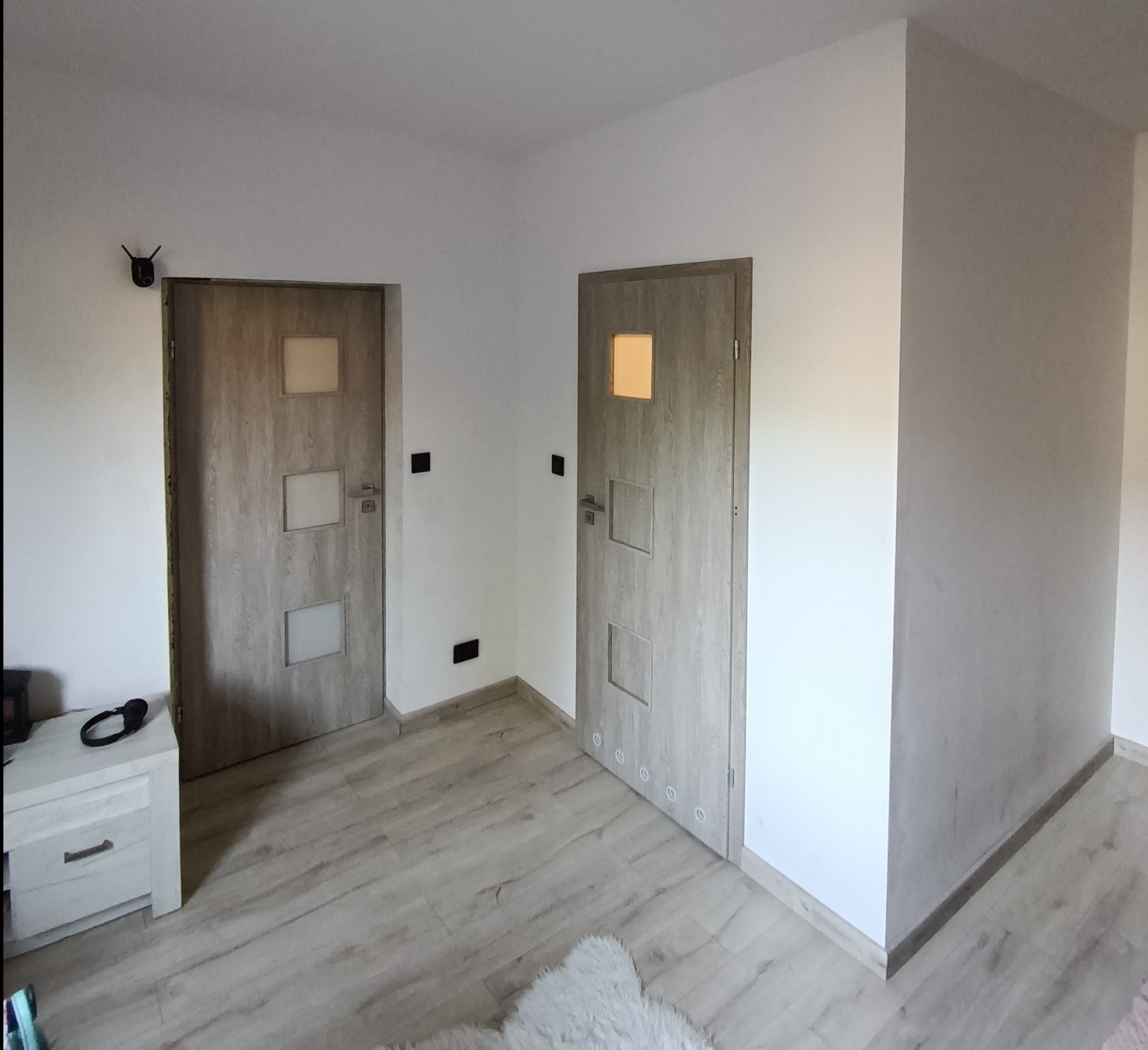 Wnętrze po remoncie: dwie drewniane drzwi z przeszkleniami, jasne ściany i podłoga z paneli. Widoczny fragment szafki nocnej i czujnik na ścianie. Minimalistyczny design.