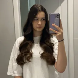 Młoda kobieta z długimi, brązowymi włosami, pozująca do selfie w łazience, trzymając telefon w fioletowym etui. Widoczna umywalka i białe drzwi w tle.