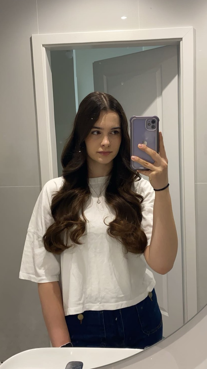 Młoda kobieta z długimi, brązowymi włosami, pozująca do selfie w łazience, trzymając telefon w fioletowym etui. Widoczna umywalka i białe drzwi w tle.