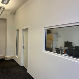 Zal-bud - Białe, minimalistyczne wnętrze biura z widokiem na pomieszczenie przez okno w ścianie. Ciemna wykładzina, białe ściany, drzwi i okno. Widoczne biurka i monitory.