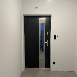 Zal-bud - Czarne drzwi wejściowe z przeszkleniem w nowym, jasnym wnętrzu. Widoczne gniazdka, szare płytki podłogowe i odbicie osoby w szybie drzwi.