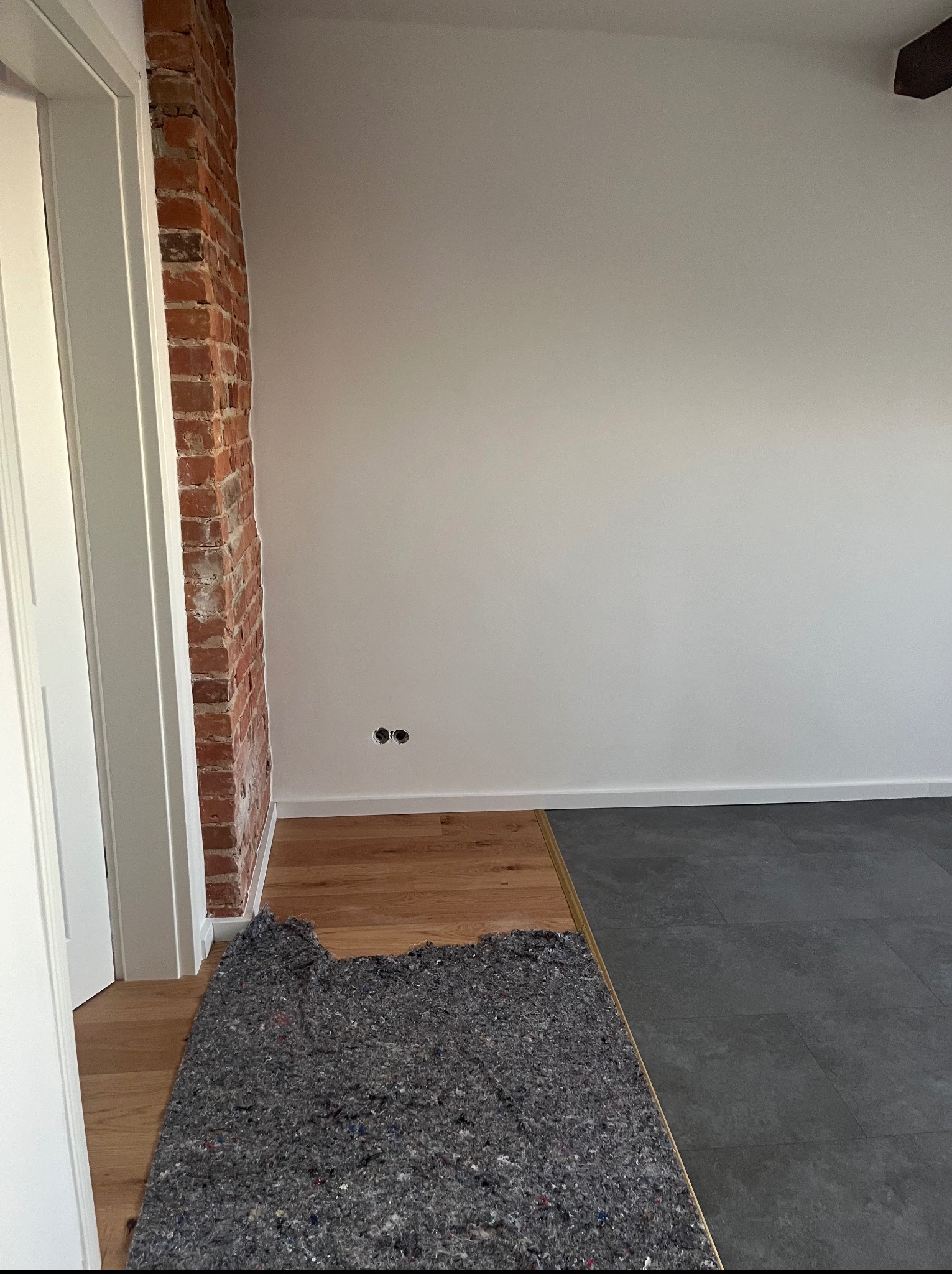 Fragment wnętrza w trakcie remontu: połączenie parkietu i płytek, zabezpieczone szarą włókniną, odsłonięta cegła. Widoczne gniazdka elektryczne i fragment sufitu.