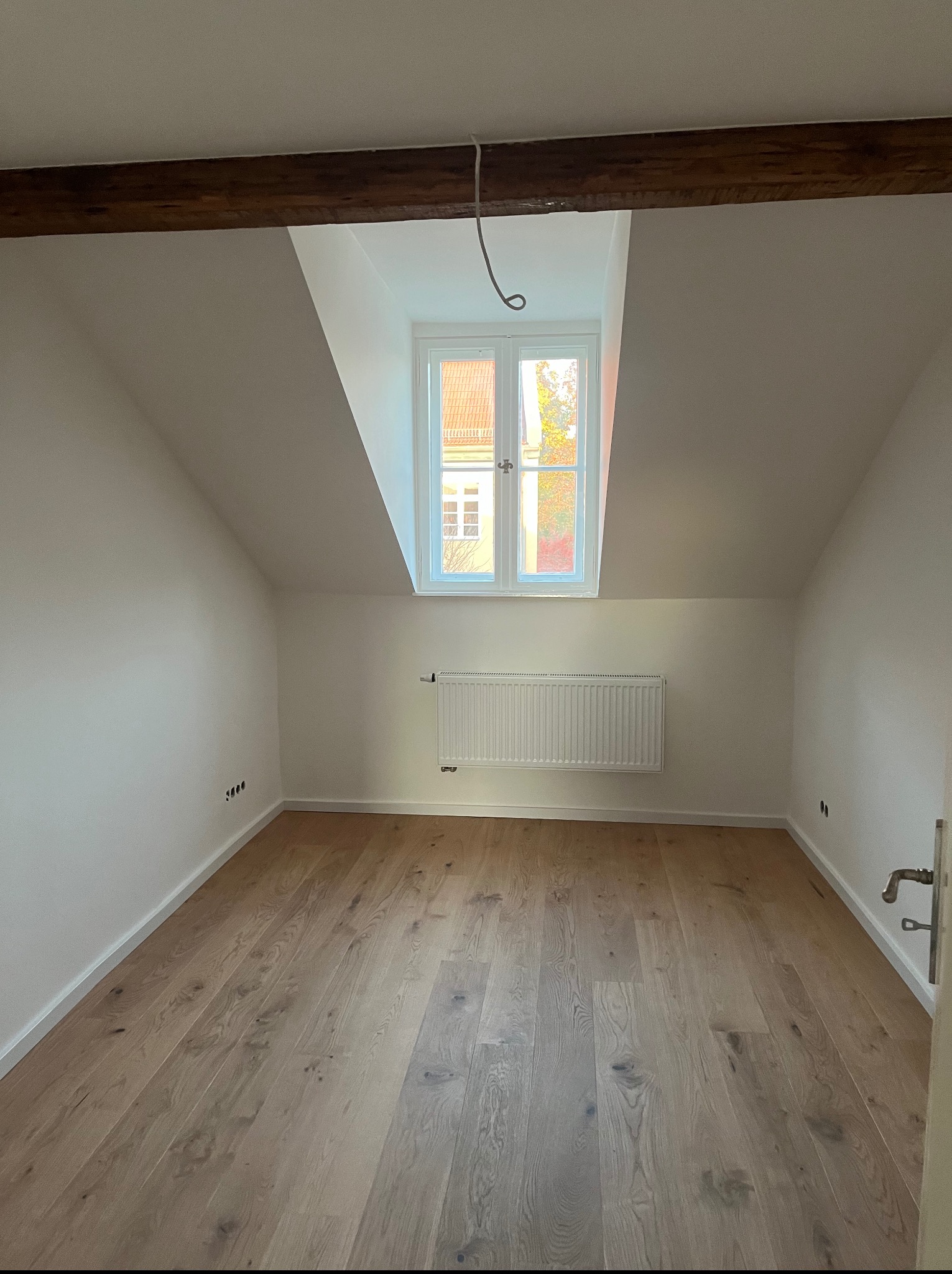 Jasne drewniane deski podłogowe w pokoju na poddaszu, białe ściany, okno dachowe z widokiem na budynki. Grzejnik na ścianie, widoczne gniazdka.