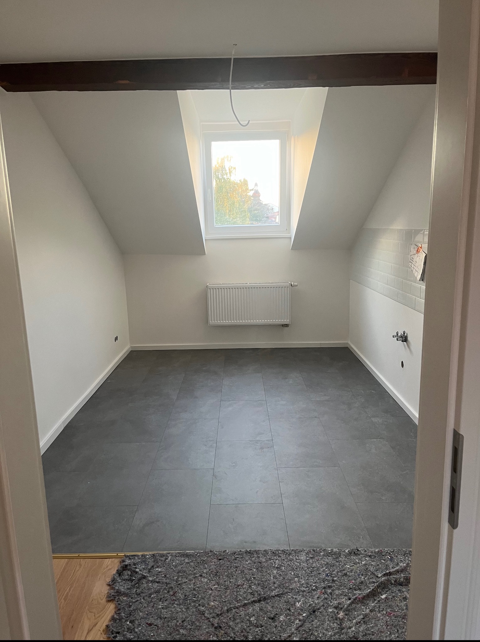 Poddasze po remoncie: szare płytki podłogowe, białe ściany, okno dachowe z widokiem na zieleń. Grzejnik na ścianie. Widok zza drzwi.
