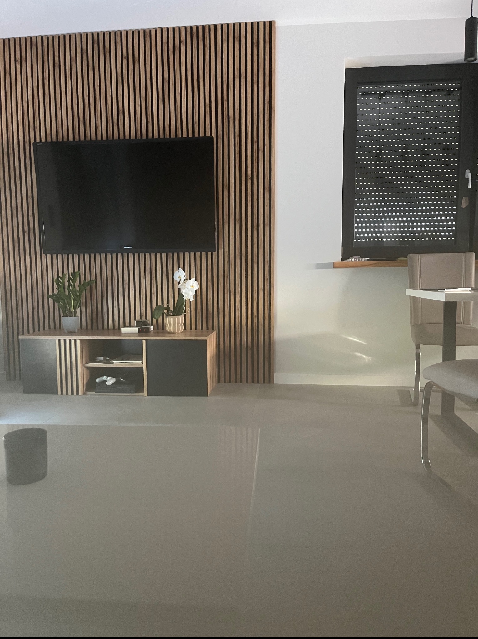 Stylowy salon w Mogilnie z drewnianą ścianą lamelową za telewizorem. Pod nim szafka RTV z roślinami. W tle okno z roletami i fragment stołu z krzesłami.