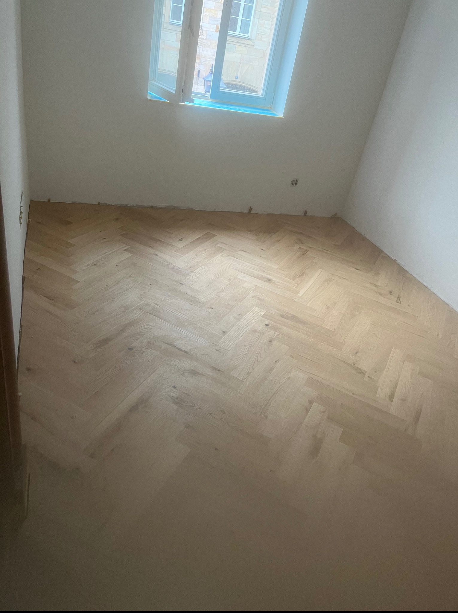 Jasny parkiet ułożony w jodełkę w pokoju z oknem. Widoczne listewki dystansowe przy ścianach, co sugeruje świeżo zakończony montaż parkietu.