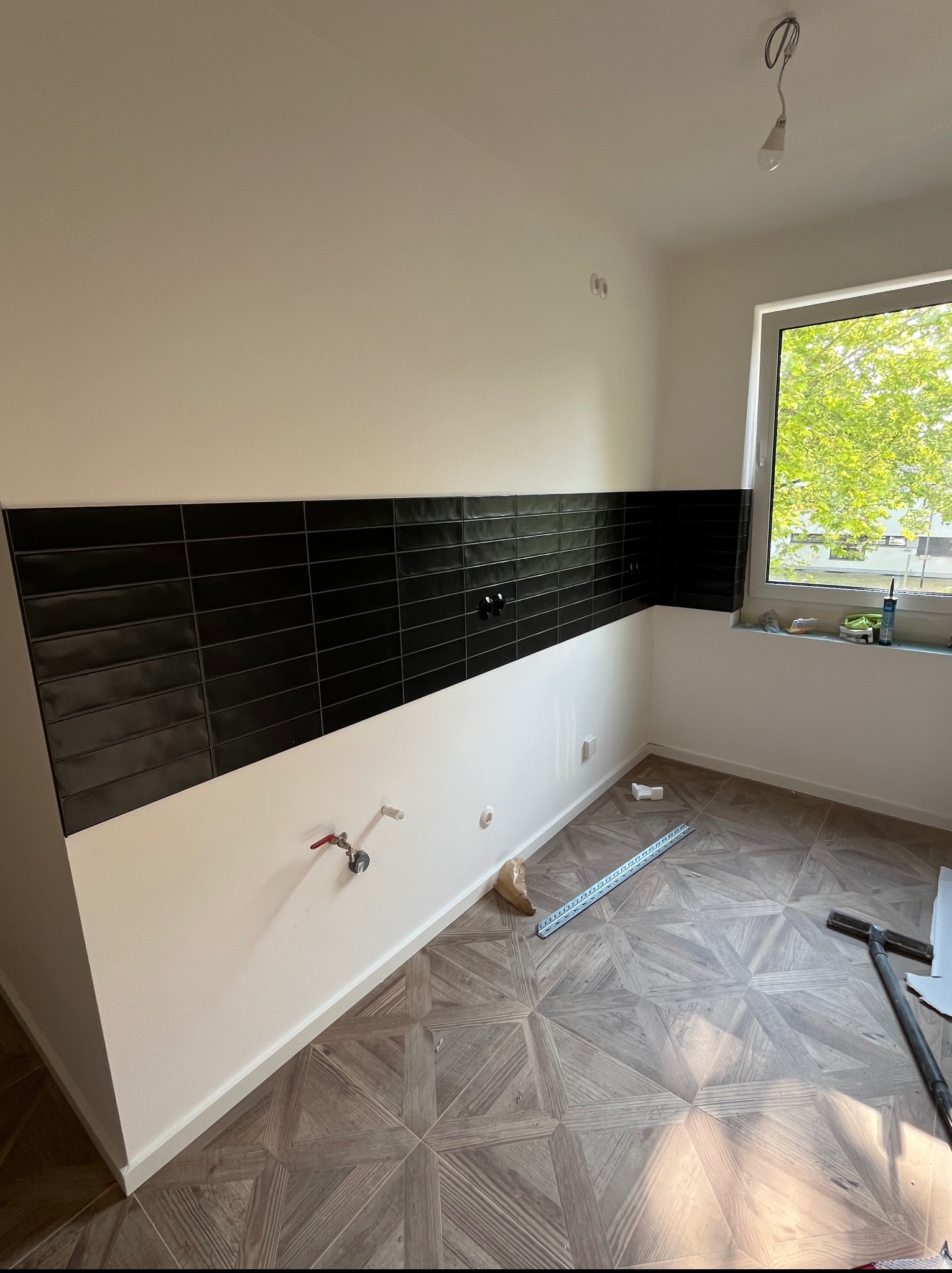 Częściowo wykończona kuchnia: czarne płytki na ścianie nad drewnianą podłogą, widoczne przyłącza wodne i okno z zielenią na zewnątrz. Etap prac wykończeniowych.