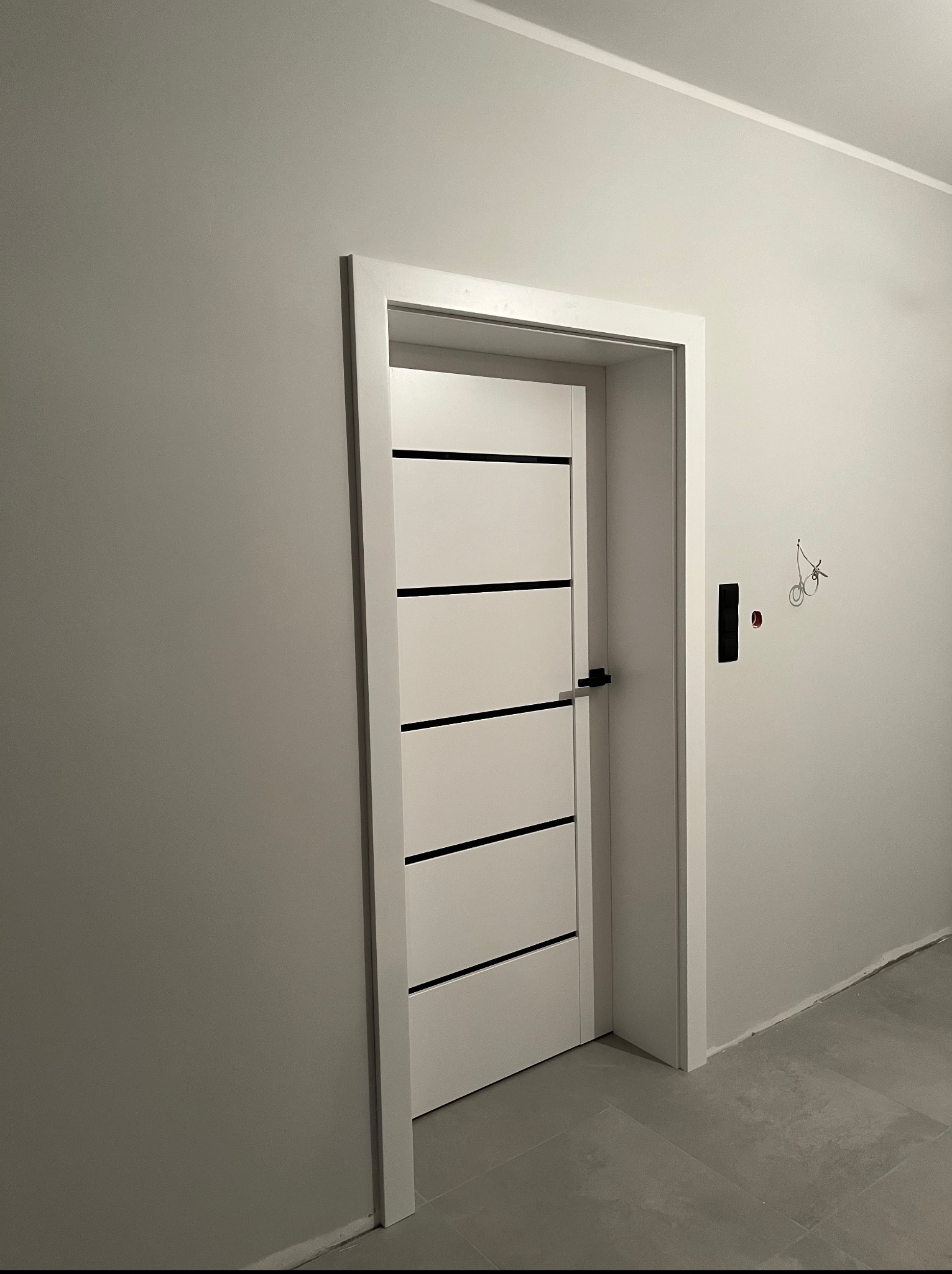 Nowoczesne, białe drzwi wewnętrzne z czarnymi poziomymi paskami, osadzone w białej ościeżnicy w szarym wnętrzu. Widoczne ślady po instalacji elektrycznej.