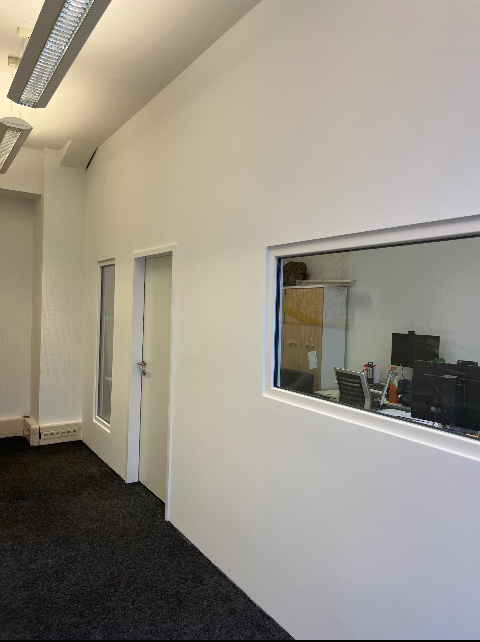 Białe, minimalistyczne wnętrze biura z widokiem na pomieszczenie przez okno w ścianie. Ciemna wykładzina, białe ściany, drzwi i okno. Widoczne biurka i monitory.