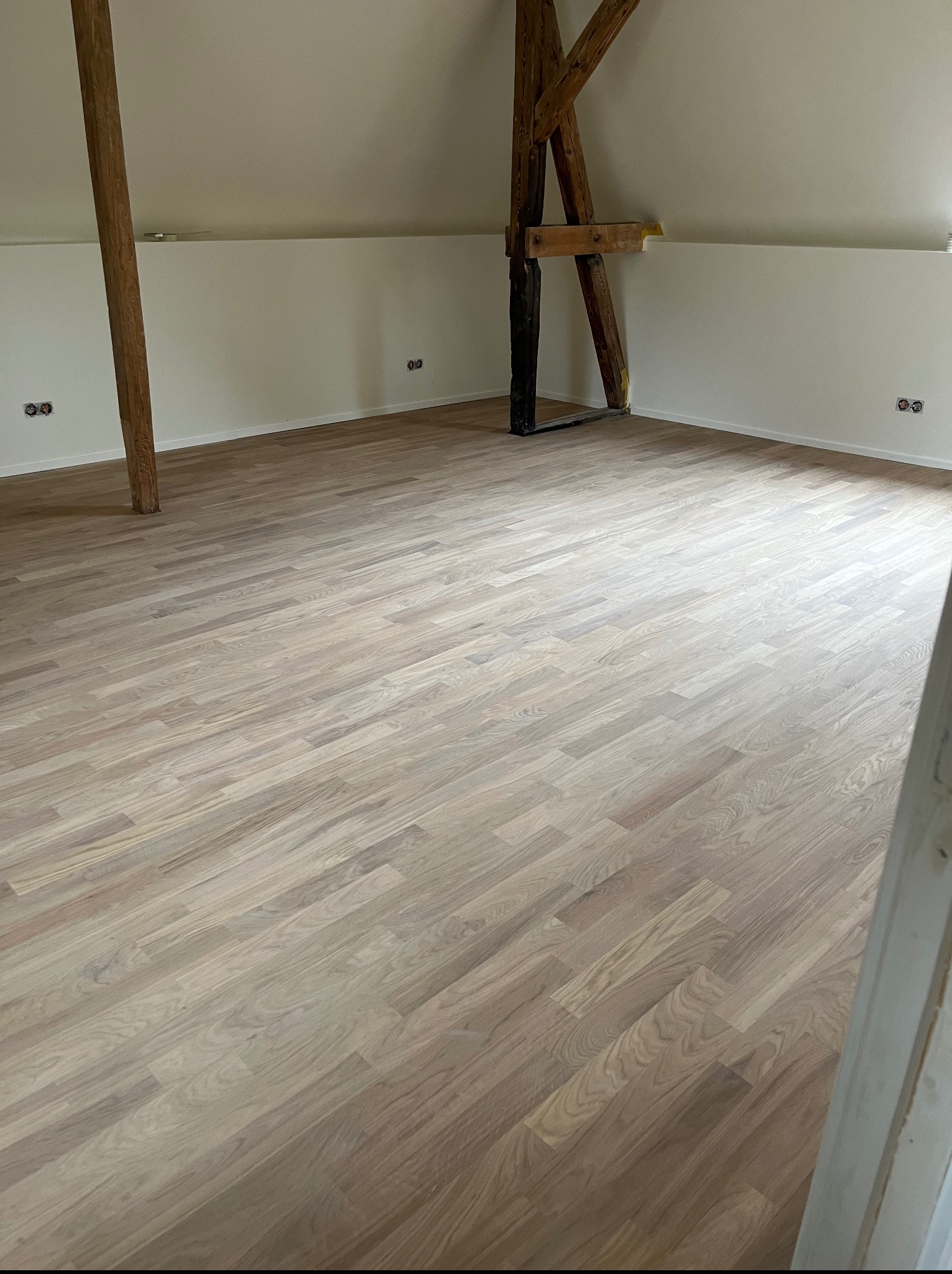 Jasny parkiet ułożony w przestronnym pokoju na poddaszu z drewnianymi belkami i białymi ścianami. Widoczne gniazdka elektryczne.