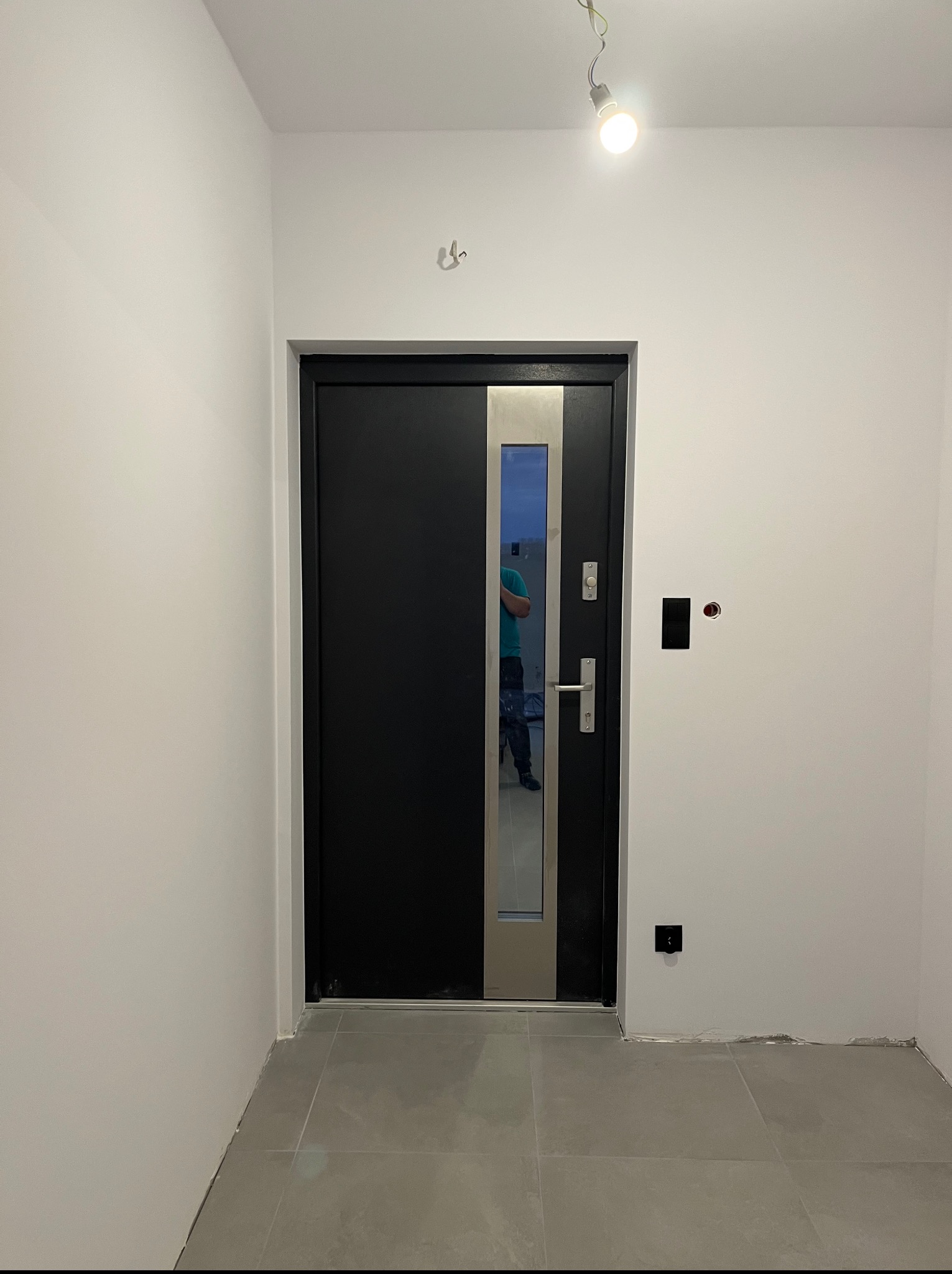 Czarne drzwi wejściowe z przeszkleniem w nowym, jasnym wnętrzu. Widoczne gniazdka, szare płytki podłogowe i odbicie osoby w szybie drzwi.