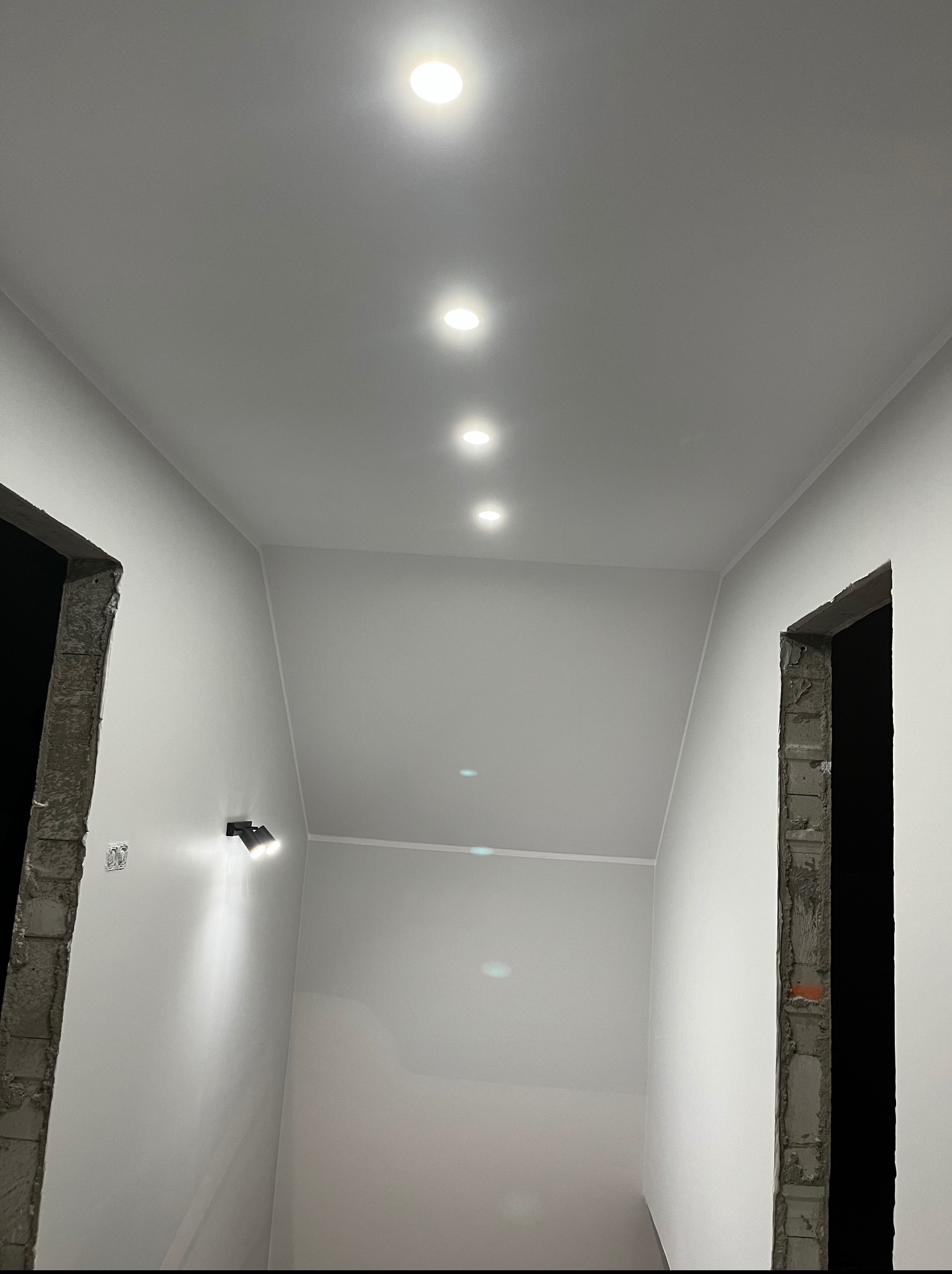 Biały korytarz w trakcie remontu z surowymi framugami drzwi i zamontowanym oświetleniem punktowym oraz lampą ścienną.