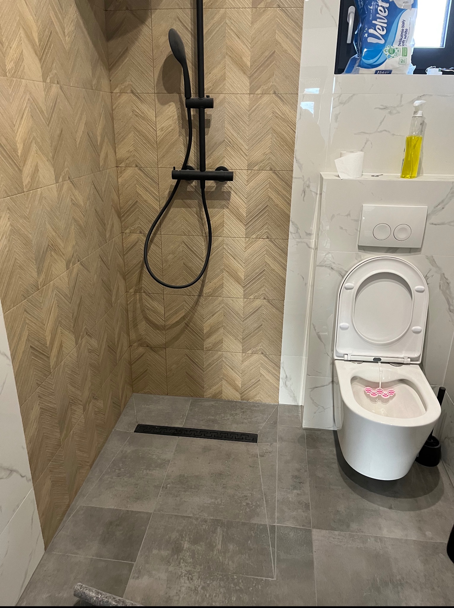 Wykończona łazienka z kabiną prysznicową typu walk-in i podwieszaną toaletą. Uwagę zwracają płytki imitujące drewno ułożone w jodełkę na ścianach kabiny.