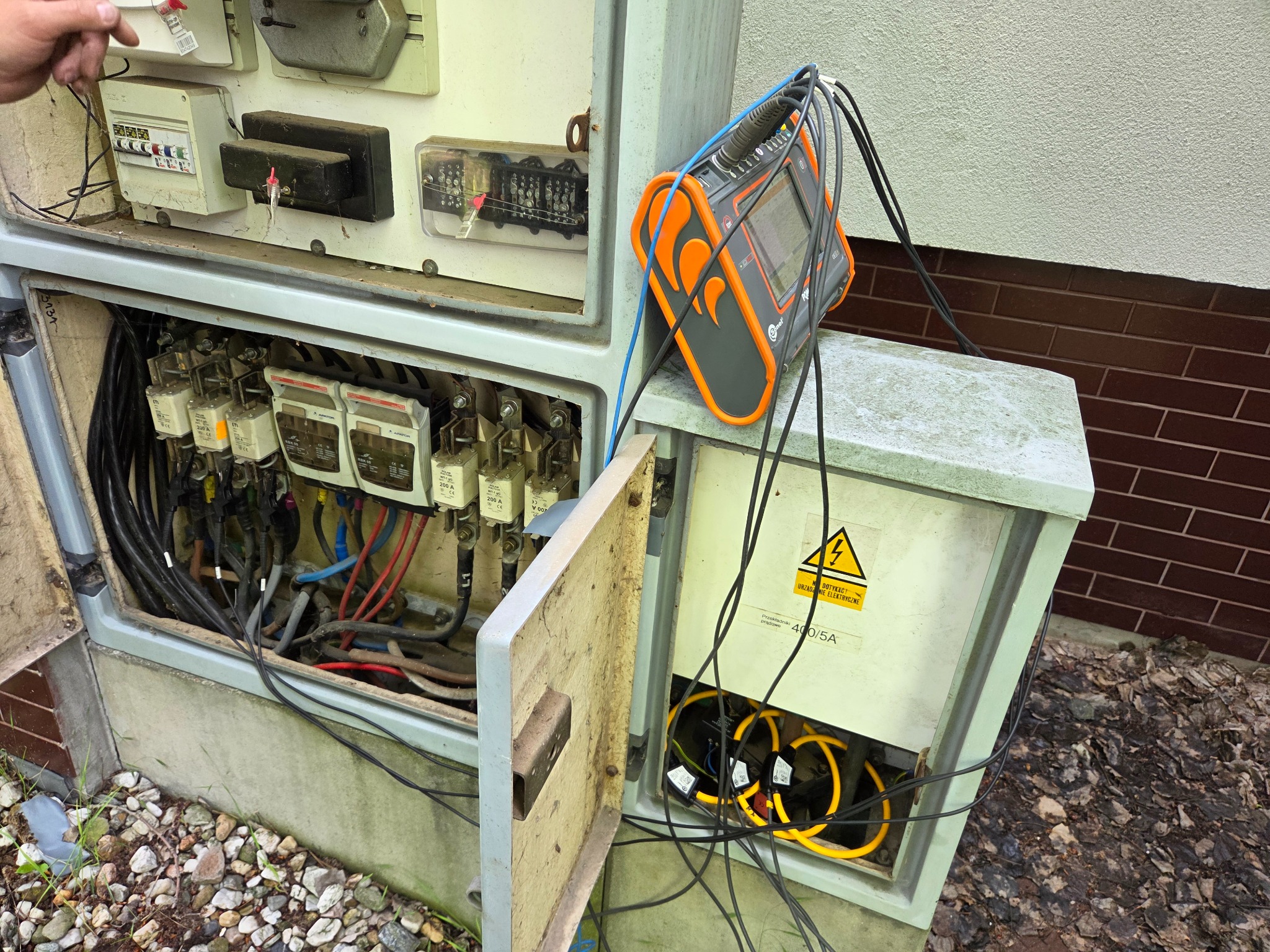 Otwarta skrzynka z licznikami energii elektrycznej i podłączonym analizatorem jakości zasilania, widoczne przewody i zabezpieczenia. Zewnętrzna instalacja elektryczna.