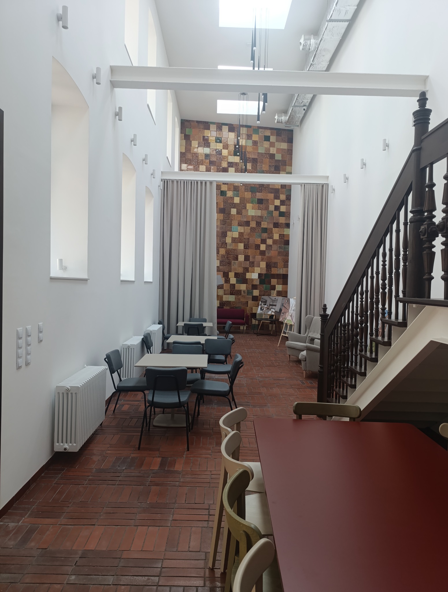 Przestronne wnętrze po remoncie w Katowicach: ceglana podłoga, białe ściany z oknami, drewniane schody i ściana z płytek. Widoczne stoliki z krzesłami i grzejniki.