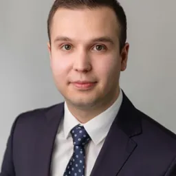 Portret młodego mężczyzny w garniturze i krawacie w kropki, z delikatnym uśmiechem, na jasnoszarym tle. Profesjonalny wizerunek.