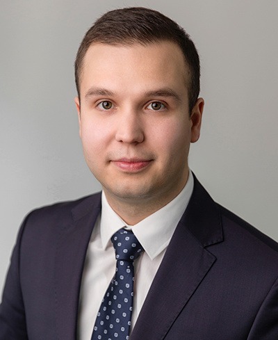 Portret młodego mężczyzny w garniturze i krawacie w kropki, z delikatnym uśmiechem, na jasnoszarym tle. Profesjonalny wizerunek.