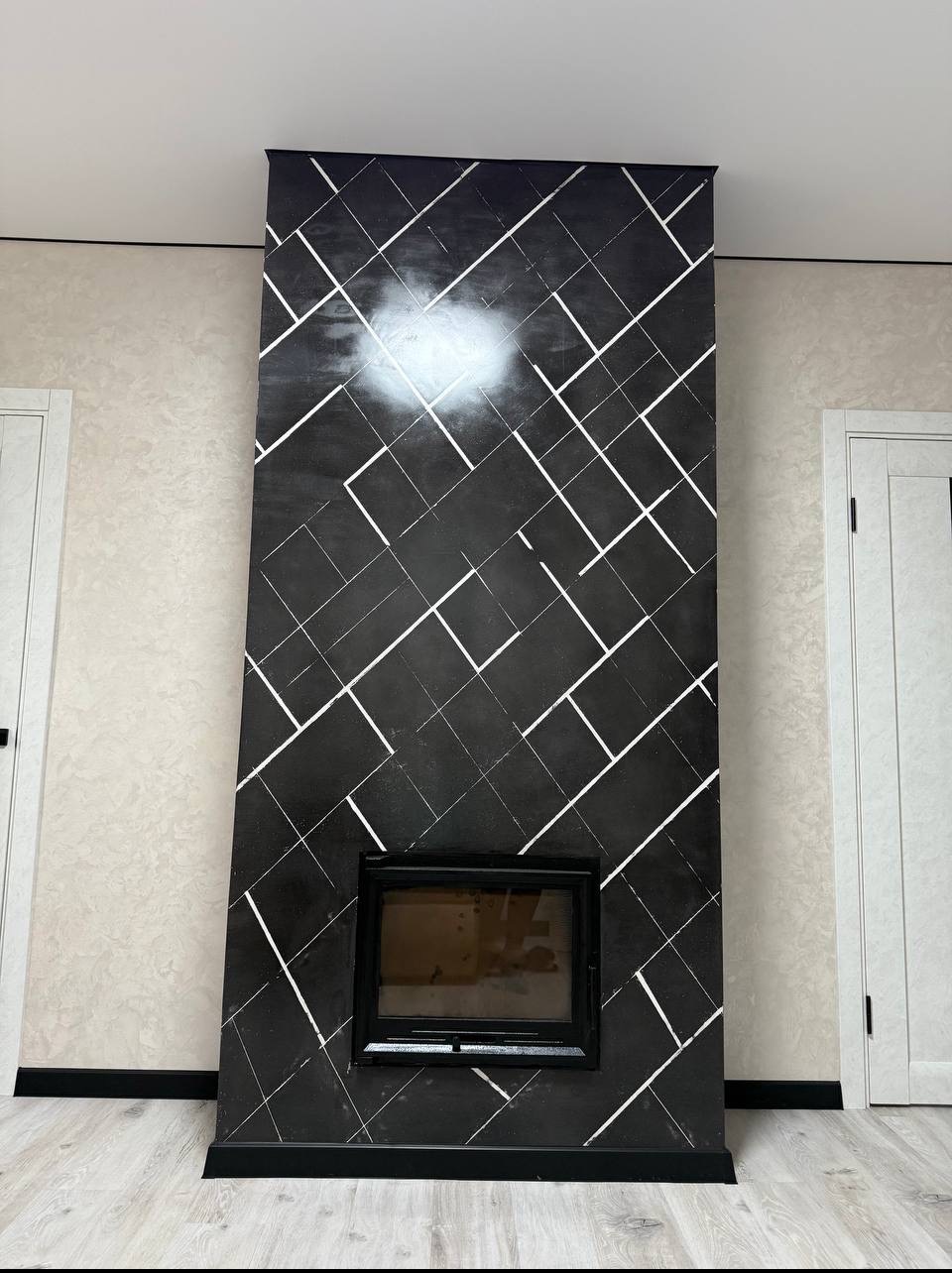 Wykończony kominek z czarną okładziną w geometryczne, białe wzory, z widocznym paleniskiem. Obok drzwi i ściana w jasnych odcieniach. Nowoczesny design.