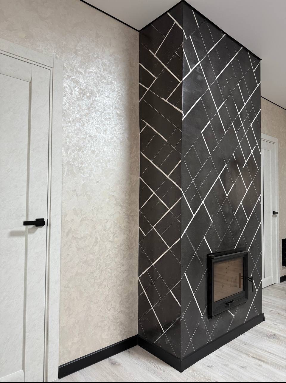 Wykończenie wnętrza: kolumna z czarnego mikrocementu z białymi liniami, wbudowany wkład kominkowy, ściana z dekoracyjną fakturą, białe drzwi z czarną klamką i czarna listwa przypodłogowa.