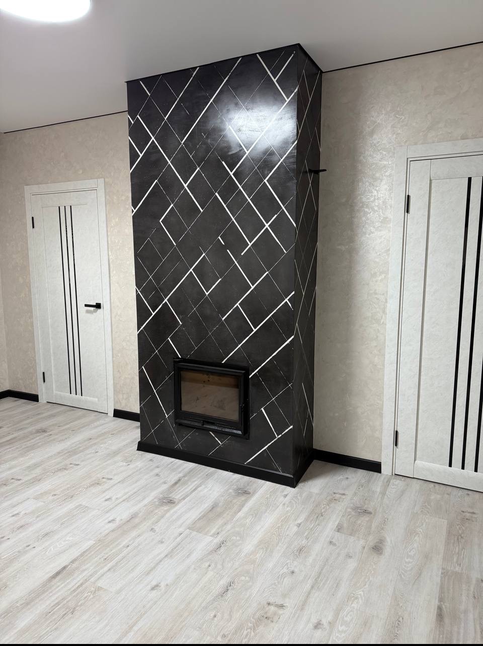 Nowoczesny kominek obłożony czarnym mikrocementem z białym geometrycznym wzorem, w jasnym wnętrzu z drewnianą podłogą i białymi drzwiami.