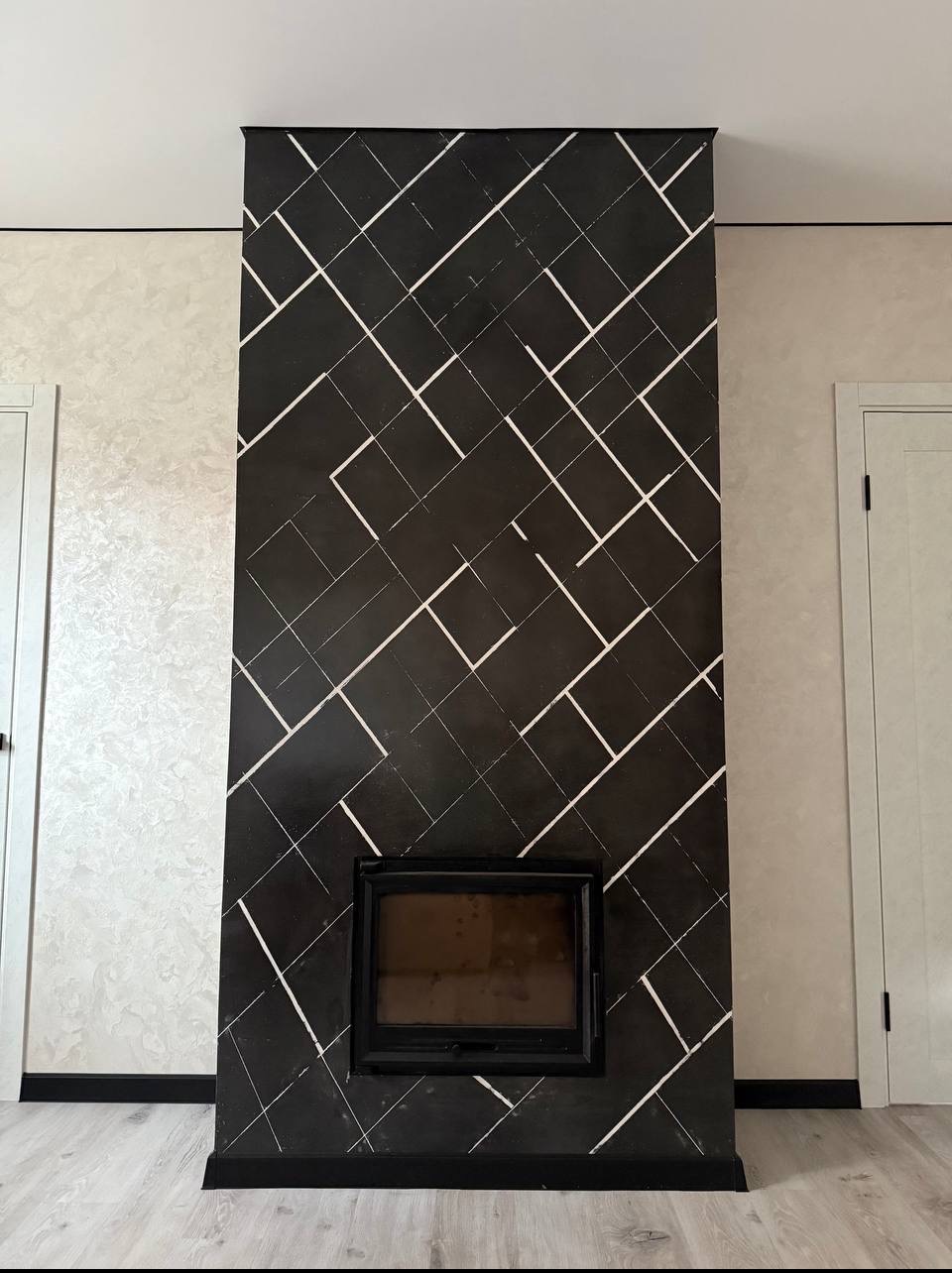 Wykończenie kominka czarnym mikrocementem z białymi liniami, obok ściana w jasnym odcieniu i białe drzwi. Nowoczesny design kominka.