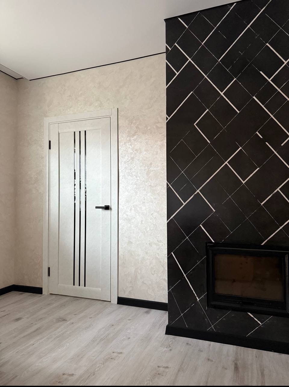 Pokój po remoncie: kremowe ściany z efektem dekoracyjnym, białe drzwi z czarnymi paskami, czarna ściana z geometrycznym wzorem i kominkiem, jasna podłoga.