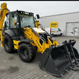 Żółta koparko-ładowarka New Holland 100D, zaparkowana na brukowanym placu przed budynkiem z logo serwisu. Widoczny lemiesz z widłami. Częściowo ośnieżona.