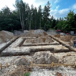Artur Kloczkowski Wolf Usłuki koparko ładowarką - Wykopane rowy pod fundamenty z wystającymi prętami zbrojeniowymi, drewniane szalunki, ziemia i las w tle, mężczyzna w oddali.