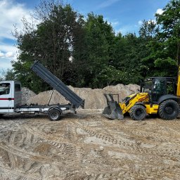 Artur Kloczkowski Wolf Usłuki koparko ładowarką - Żółta koparko-ładowarka New Holland i biała wywrotka na piaszczystym terenie, z kopcem piasku w tle i zielenią drzew. Ślady opon na piasku.
