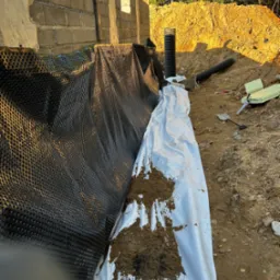 Ocieplenie fundamentów z użyciem folii kubełkowej, białej geowłókniny i rury drenarskiej. Widoczny wykop i narzędzia.