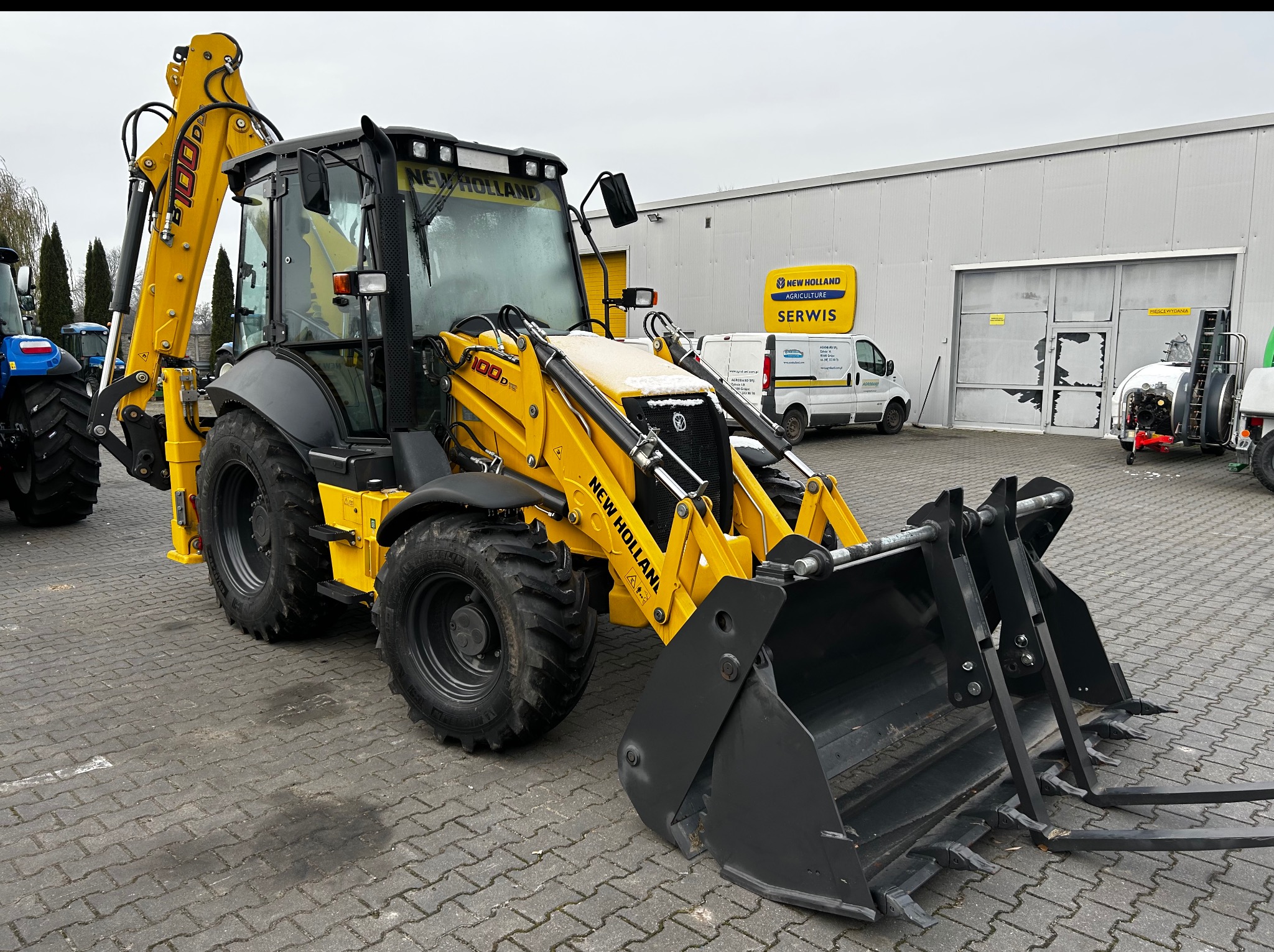 Żółta koparko-ładowarka New Holland 100D, zaparkowana na brukowanym placu przed budynkiem z logo serwisu. Widoczny lemiesz z widłami. Częściowo ośnieżona.
