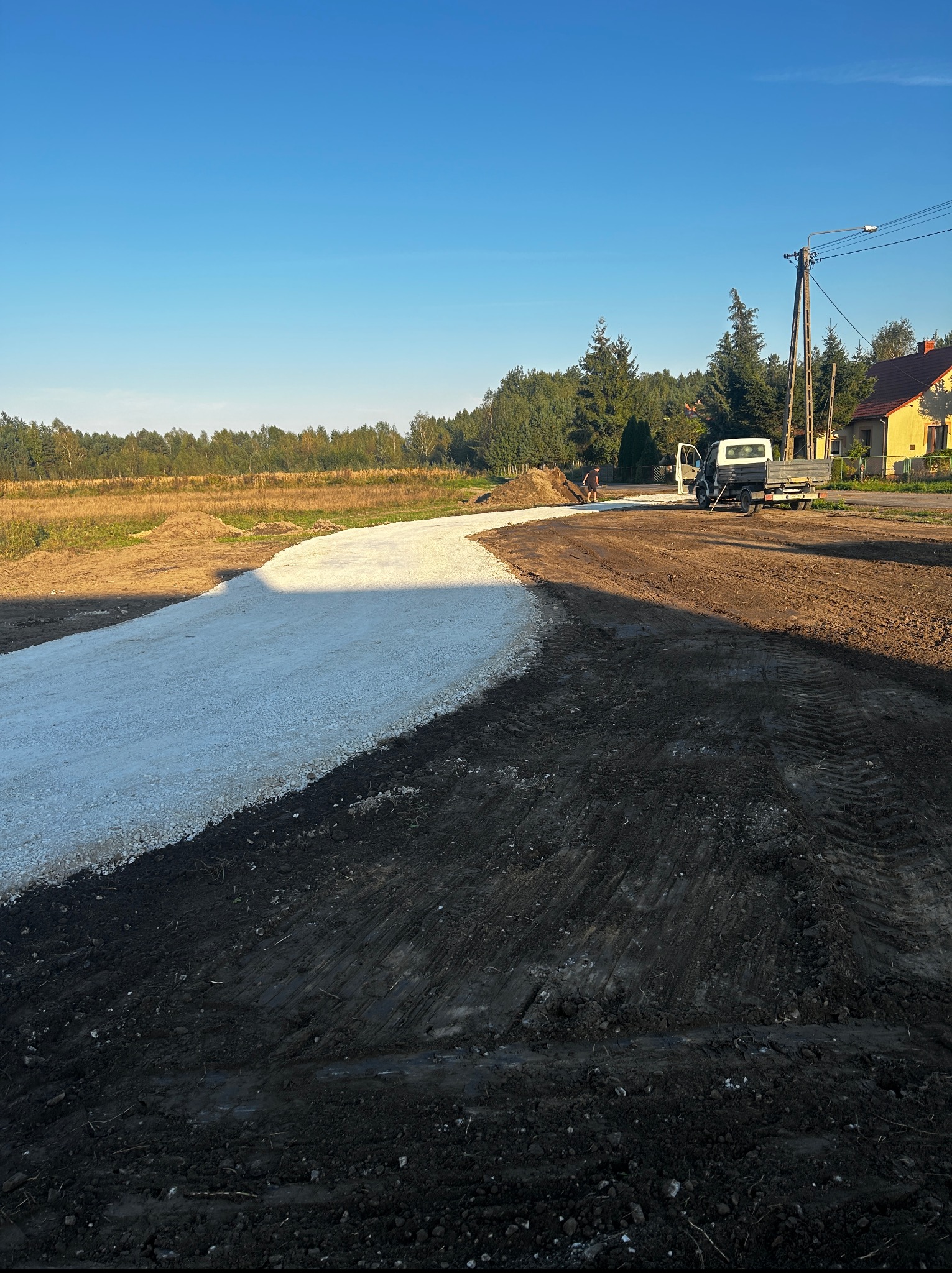 Niwelacja terenu pod drogę dojazdową: wyrównana, jasna nawierzchnia z kruszywa kontrastuje z ciemną ziemią. W tle dom, drzewa, błękitne niebo.
