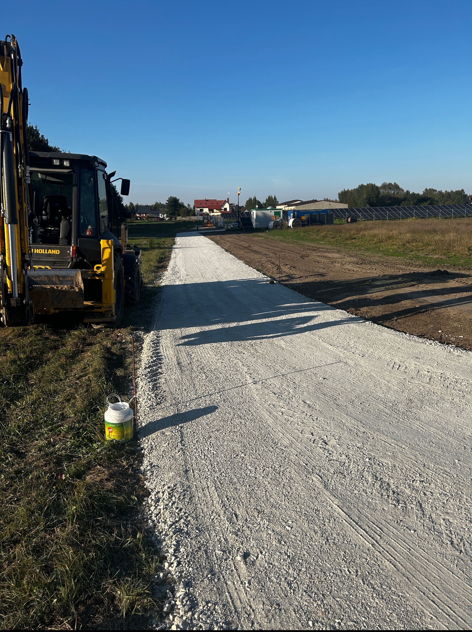 Żółta koparko-ładowarka New Holland obok wyrównanej, szarej drogi gruntowej na tle błękitnego nieba i zabudowań gospodarczych w oddali.