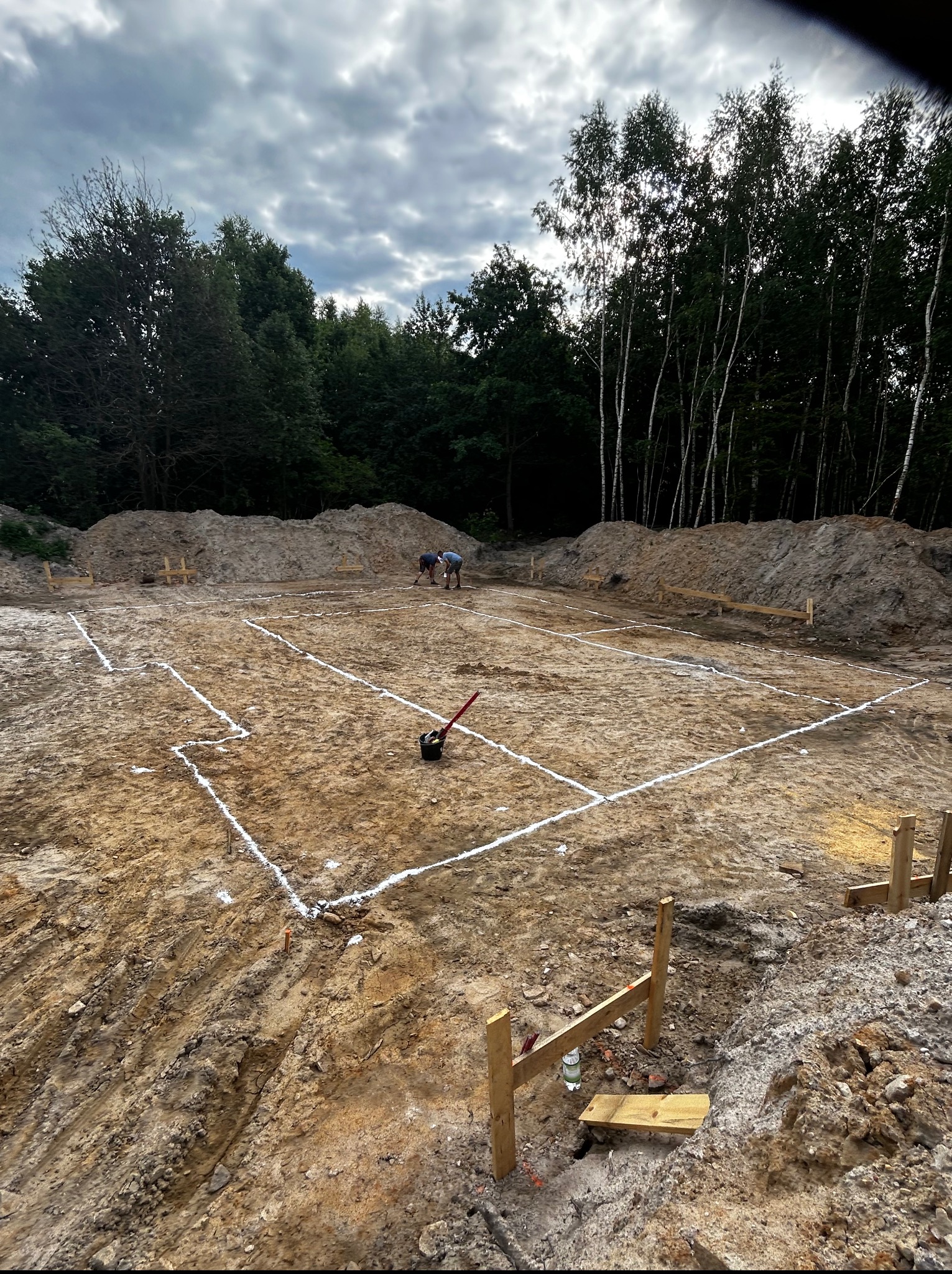 Wytyczanie fundamentów: dwóch pracowników nanosi białe linie na przygotowany teren pod budowę, widoczne narzędzia i drewniane paliki. W tle las i pochmurne niebo.
