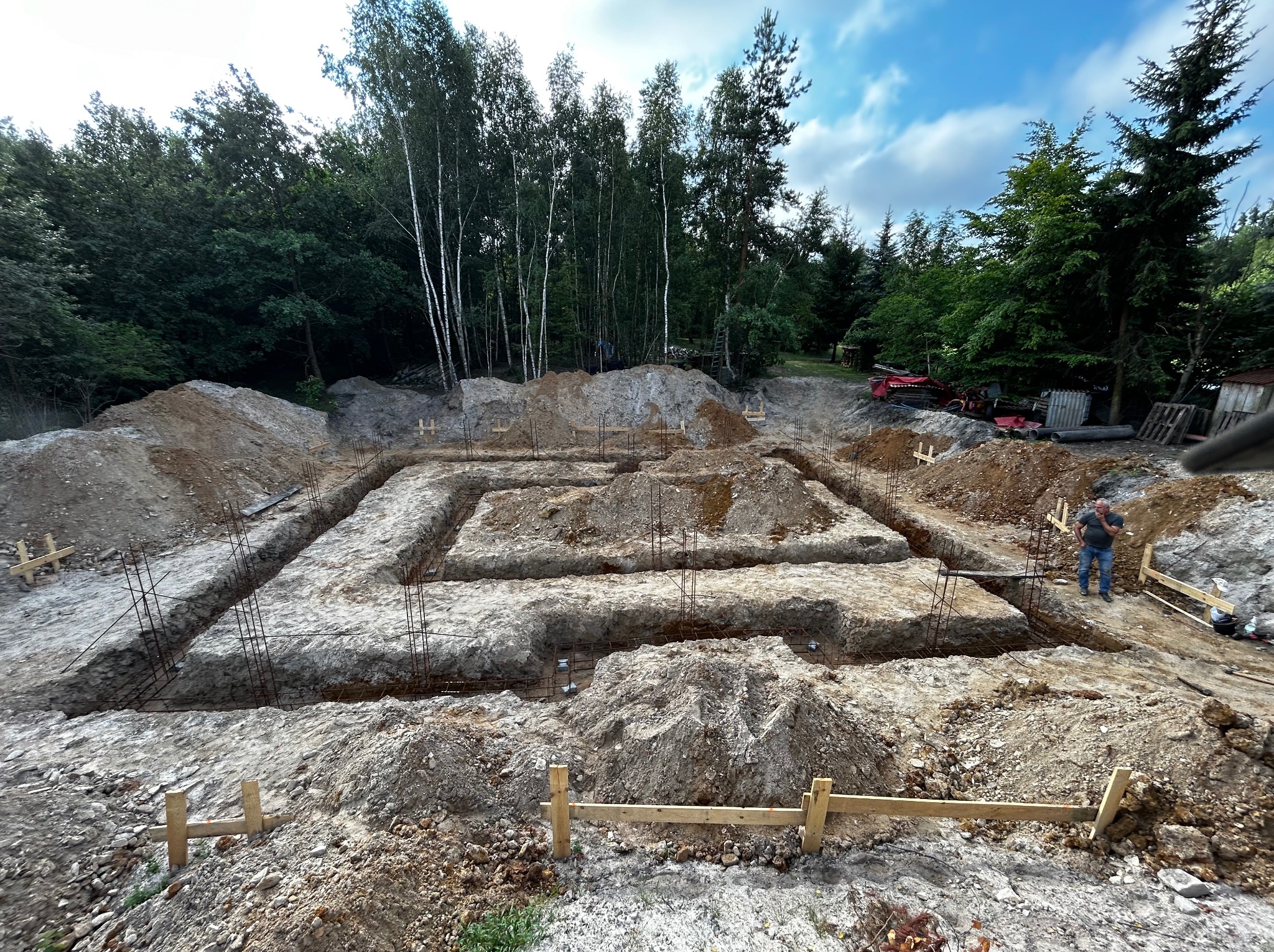 Wykopane rowy pod fundamenty z wystającymi prętami zbrojeniowymi, drewniane szalunki, ziemia i las w tle, mężczyzna w oddali.
