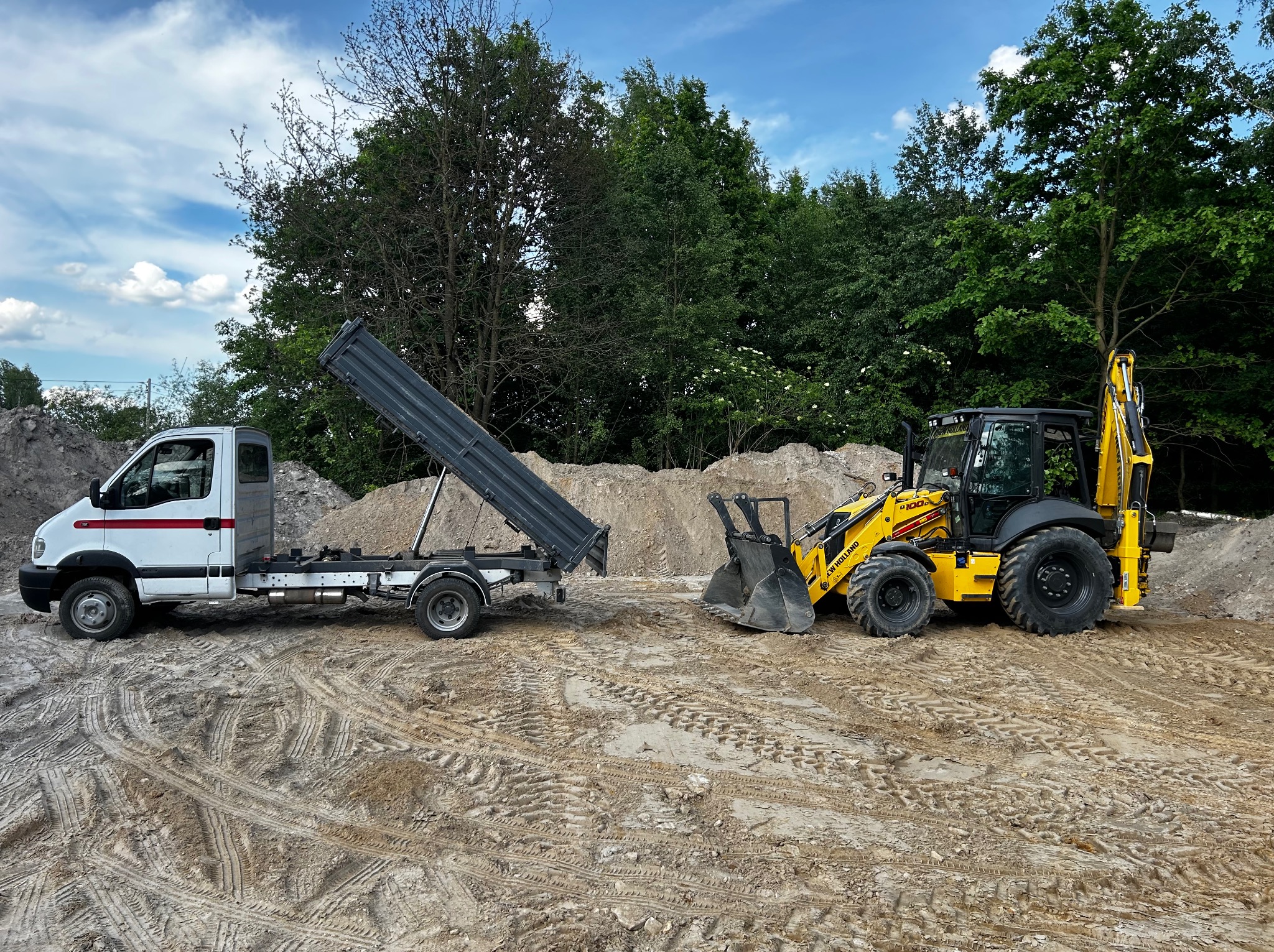 Żółta koparko-ładowarka New Holland i biała wywrotka na piaszczystym terenie, z kopcem piasku w tle i zielenią drzew. Ślady opon na piasku.