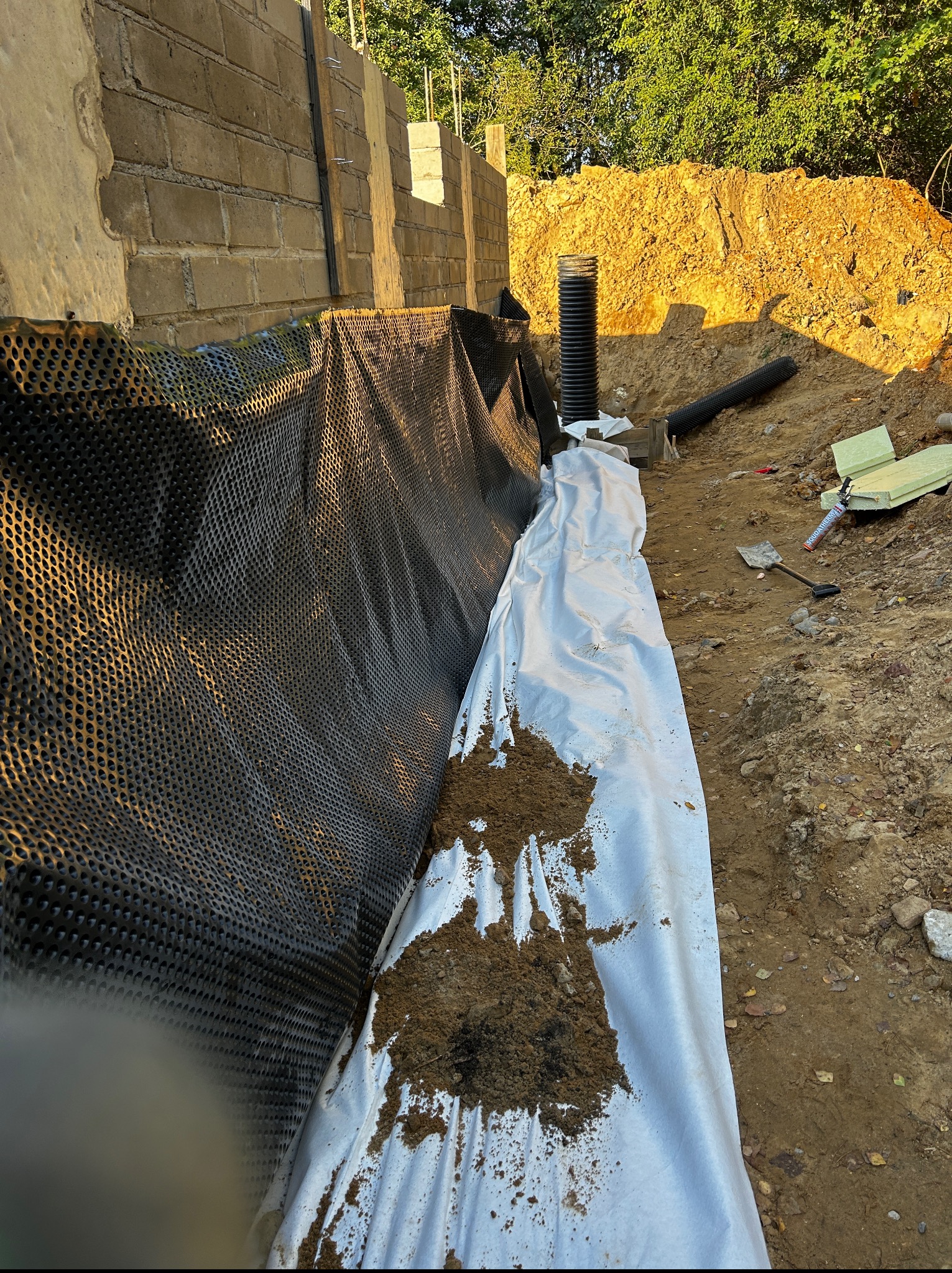 Ocieplenie fundamentów z użyciem folii kubełkowej, białej geowłókniny i rury drenarskiej. Widoczny wykop i narzędzia.