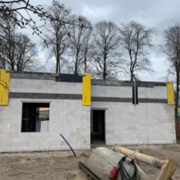 Wznoszenie murów domu z betonu komórkowego z widocznym oknem i otworem drzwiowym. Szalunki pionowe w kolorze żółtym. Na pierwszym planie betoniarka.